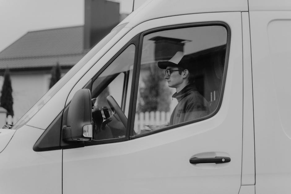 A man wearing glasses and a cap sits inside the cab of a white van, visible through the front passenger window during daylight hours. The van is parked outside a residential property, with houses and trees in the background. Inside the vehicle, the driver appears focused, with a dashboard and steering wheel visible. The side mirror extends outward, and the van's door handle is seen on the exterior. This scene depicts a professional removals process, likely part of home relocation or furniture transport services offered by Kingston Upon Thames Removals, highlighting the loading or journey phase of house removals in the KT1 and KT2 postal code areas.