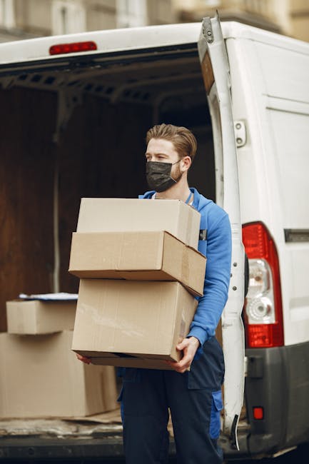 A young man with light skin, wearing a black face mask and a blue hoodie, is standing next to an open white moving van. He is carrying three cardboard boxes stacked on top of each other, with the top box slightly smaller than the middle and bottom boxes. Inside the van, visible in the background, are additional cardboard boxes and packing materials arranged on the van's floor and side shelves. The scene is set on a street or driveway, with natural daylight illuminating the area, and some blurred building details in the background. This image captures the process of home relocation, with furniture and packing boxes being loaded or unloaded as part of a professional removals service managed by Kingston Upon Thames Removals, which offers house moving and furniture transport services in the KT1 and KT2 areas.