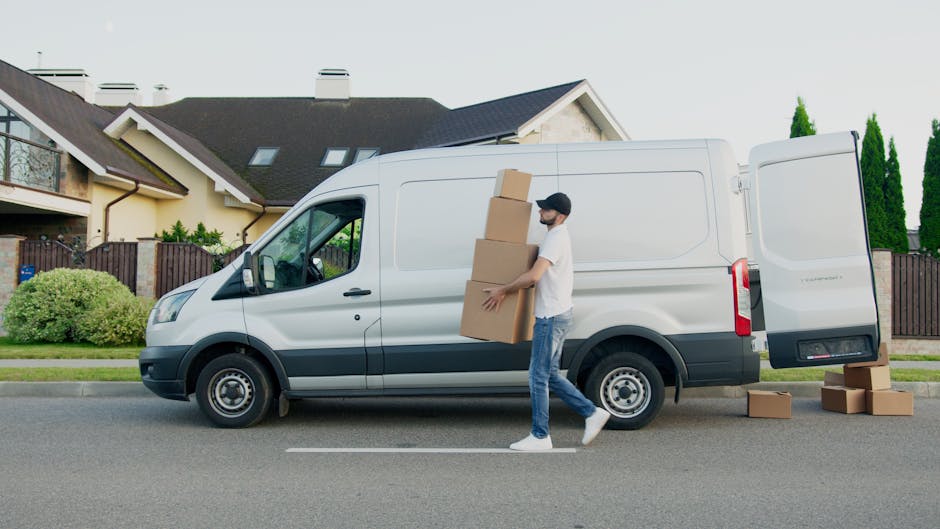 A man wearing glasses and a cap sits inside the cab of a white van, visible through the front passenger window during daylight hours. The van is parked outside a residential property, with houses and trees in the background. Inside the vehicle, the driver appears focused, with a dashboard and steering wheel visible. The side mirror extends outward, and the van's door handle is seen on the exterior. This scene depicts a professional removals process, likely part of home relocation or furniture transport services offered by Kingston Upon Thames Removals, highlighting the loading or journey phase of house removals in the KT1 and KT2 postal code areas.