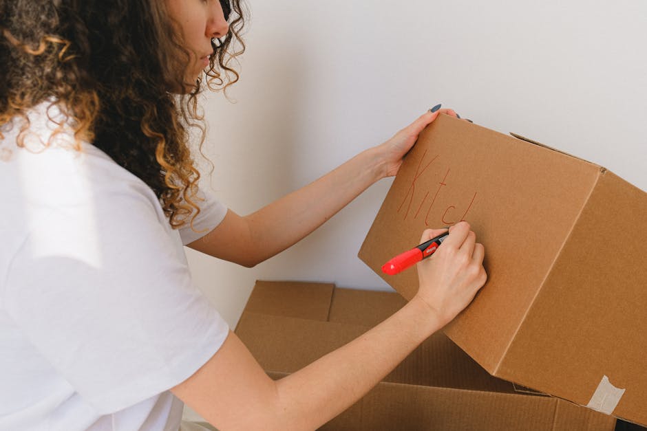 A woman with curly hair, wearing a white t-shirt, is in the process of packing for a home relocation inside a room with neutral-colored walls. She is holding a brown cardboard box labeled 'Kitchen' in red marker, preparing it for moving. The woman is using a black and red marker to write on the box, ensuring proper labeling for organized furniture transport. Surrounding her are additional empty cardboard boxes, some stacked and others on the floor, ready for packing household items. The scene captures the loading process of packing and moving, with natural lighting illuminating the space, reflecting standard packing practices during house removals. This image emphasizes careful packing, appropriate use of cardboard boxes, and labeling, aligned with professional removals services offered by Kingston Upon Thames Removals, supporting the topic of avoiding hidden moving costs and ensuring an efficient home relocation process.