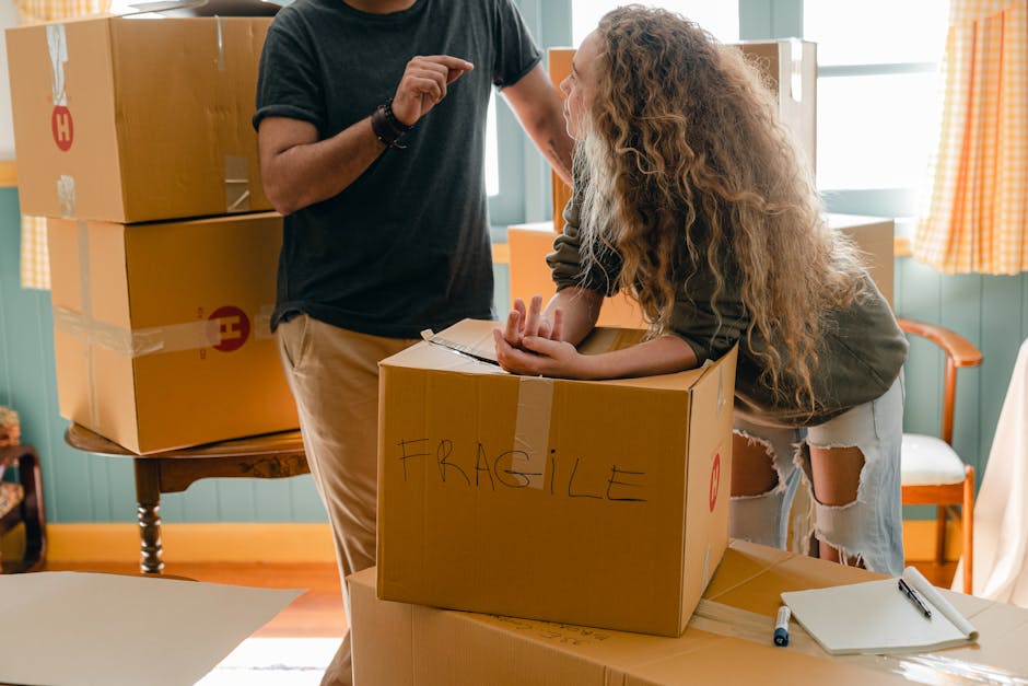 A person wearing a black T-shirt and beige trousers is assisting a woman with long, curly hair and light-colored, ripped jeans in packing fragile items into a cardboard box labeled 'FRAGILE' during a home relocation process. The woman is leaning over the box with her hands resting on the edge, while the man appears to be explaining or giving instructions, with his hand raised. The scene is set inside a room with blue-green walls, orange and white checked curtains on a window, and a wooden table with additional packed boxes stacked on top. The boxed items are secured with clear plastic wrap, indicative of careful packing and transport, as part of a furniture move organized by Kingston Upon Thames Removals. A white notepad and a pen are on the table nearby, and the lighting suggests natural daylight, highlighting the ongoing packing and loading process typical of professional removals and home relocation services, with an emphasis on proper packing techniques to avoid hidden costs in moving.