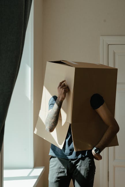 A woman with curly hair, wearing a white t-shirt, is in the process of packing for a home relocation inside a room with neutral-colored walls. She is holding a brown cardboard box labeled 'Kitchen' in red marker, preparing it for moving. The woman is using a black and red marker to write on the box, ensuring proper labeling for organized furniture transport. Surrounding her are additional empty cardboard boxes, some stacked and others on the floor, ready for packing household items. The scene captures the loading process of packing and moving, with natural lighting illuminating the space, reflecting standard packing practices during house removals. This image emphasizes careful packing, appropriate use of cardboard boxes, and labeling, aligned with professional removals services offered by Kingston Upon Thames Removals, supporting the topic of avoiding hidden moving costs and ensuring an efficient home relocation process.