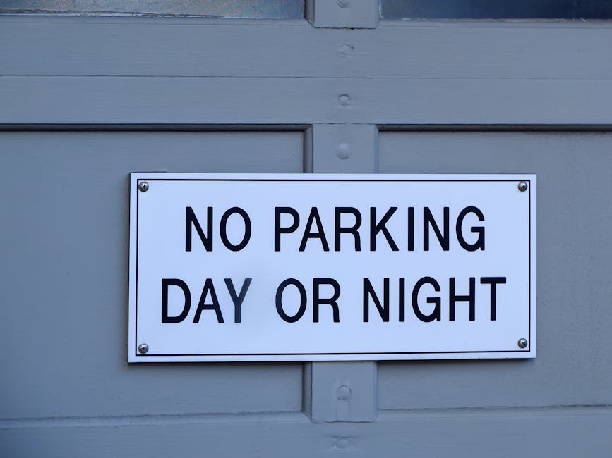 A rectangular white metal sign with black capitalized text reading 'NO PARKING DAY OR NIGHT,' mounted on a light grey wooden building structure. The sign is secured with four screws at each corner and positioned above a small, protruding horizontal wooden panel. Inside the scene, there is no visible traffic, vehicles, or people, just the sign attached to the building exterior. The lighting appears natural, indicating daytime conditions. This image relates to house removals and loading processes, illustrating parking restrictions that may affect furniture transport and home relocation logistics, and is featured on a webpage about Kingston Council parking rules for removals and loading, associated with Kingston Upon Thames Removals.