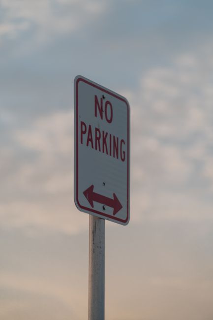 A vertical metal post supporting a rectangular 'No Parking' sign with a white background and bold red lettering, along with a double-headed red arrow at the bottom indicating no parking is allowed in both directions. The sign is positioned on a pavement outside a residential property, with part of a house visible behind it, including a window and doorframe. In front of the house, there are stacked cardboard boxes and moving blankets lying on the ground, along with a trolley or hand truck partially visible, used for moving furniture or boxes. The scene appears to be part of a home relocation or furniture transport process, with natural daylight and a cloudy sky overhead. Kingston Upon Thames Removals' service related to house removals and loading procedures is indirectly supported by the contextual setting of moving materials and the parking restriction sign.