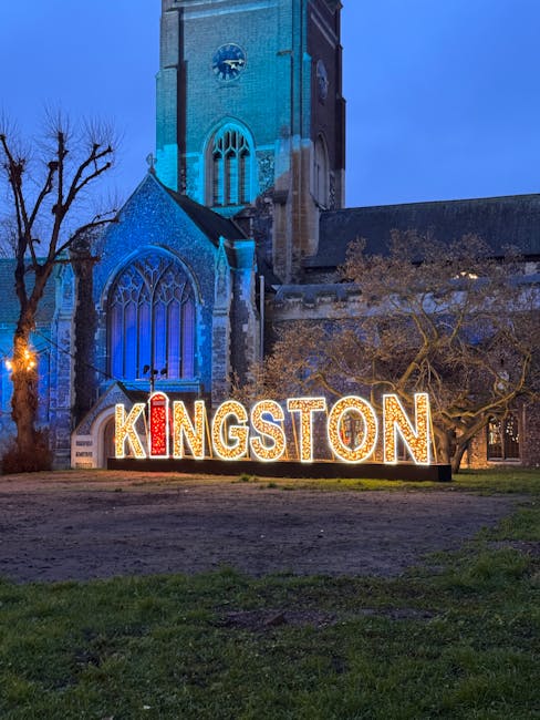 Nighttime view of a historic stone church with large stained glass windows, illuminated with blue lighting on the facade. In front of the church, there is a large, illuminated sign spelling 'KINGSTON' with individual letters in red and yellow lights, placed on the grassed area. To the right, a tree with sparse branches and some foliage is visible, while on the left, a leafless tree and exterior lighting near the church add to the scene. This setting reflects a prominent Kingston location, with no visible furniture, boxes, or moving equipment in the scene, suggesting the image captures a landmark rather than active loading or packing processes related to house removals by Kingston Upon Thames Removals.