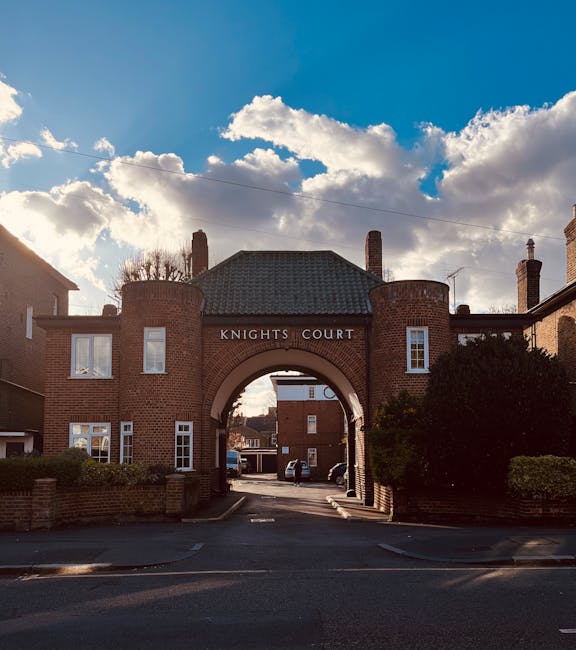 A brick residential building complex named Knights Court, with a central archway entrance leading into a parking area, is visible in this outdoor scene. The structure has a traditional design with two floors, white-framed windows, and a tiled roof. Several cars are parked inside the courtyard beyond the arch, and the sky above is partly cloudy with patches of blue visible through the clouds. The building is flanked by greenery and bushes, and the pavement in front of the entrance appears to be used for loading and unloading during a home relocation process. This scene relates to house removals and furniture transport, consistent with services provided by Kingston Upon Thames Removals, as they assist with packing, loading, and moving household items in the area near Kingston Station.