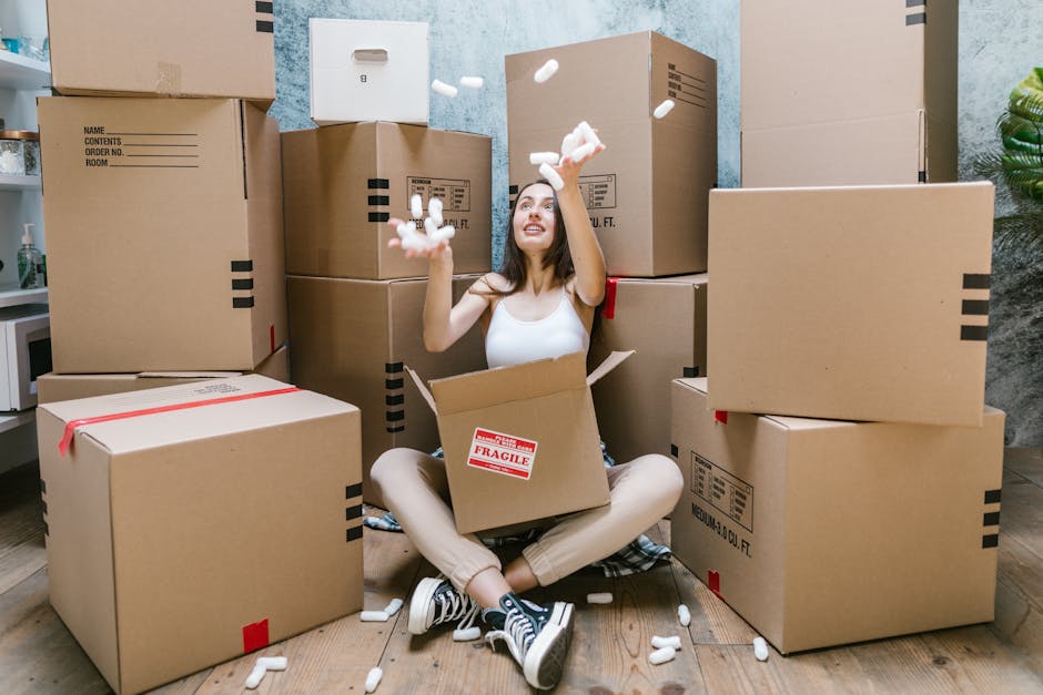 A young woman with long dark hair sitting cross-legged on a wooden floor surrounded by numerous large cardboard moving boxes, some labeled as fragile and securely sealed with red tape. She is inside a