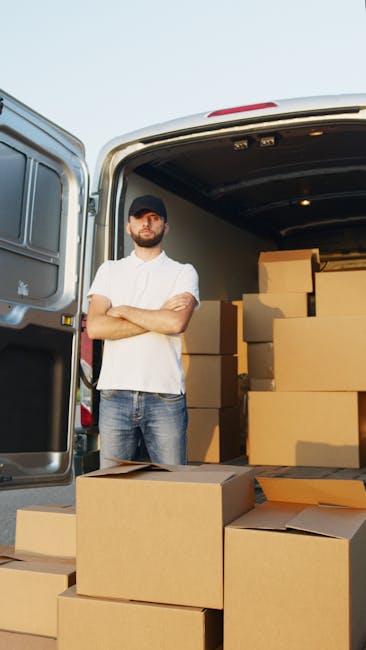 A man wearing a white shirt, dark cap, and jeans stands with arms crossed in front of an open van during a home relocation, with numerous stacked cardboard boxes inside and outside the vehicle. The boxes vary in size, some are open with flaps folded back, while others are sealed or partially packed, prepared for furniture transport and moving logistics. The van is parked outdoors in a paved area, with the rear doors fully open, revealing the interior illuminated by a small light. Trolley straps and moving blankets are visible inside the van, indicating an organized loading process related to professional removals. The environment suggests an active packing and loading stage, typical of a same day removals service provided by Kingston Upon Thames Removals, as seen on the company’s website, [DOMAIN], with the page titled ‘Same day removals Kingston Upon Thames availability and rates’, emphasizing efficient and prompt transfer of household belongings.