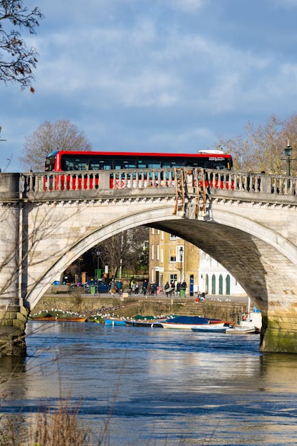 A red and white bus is traveling across a stone arch bridge over a river in a city. The bridge features decorative balustrades and a small ledge along the edges. Beneath the bridge, the river reflects the sky and surrounding structures with boats moored along the bank. In the background, there are multi-storey buildings and leafless trees indicating a cooler season. The scene is outdoors during daylight with partly cloudy skies. This setting showcases a typical urban environment suitable for a home relocation or moving service, as provided by Kingston Upon Thames Removals, highlighting the transportation and logistic aspects of furniture transport during packing and moving processes.