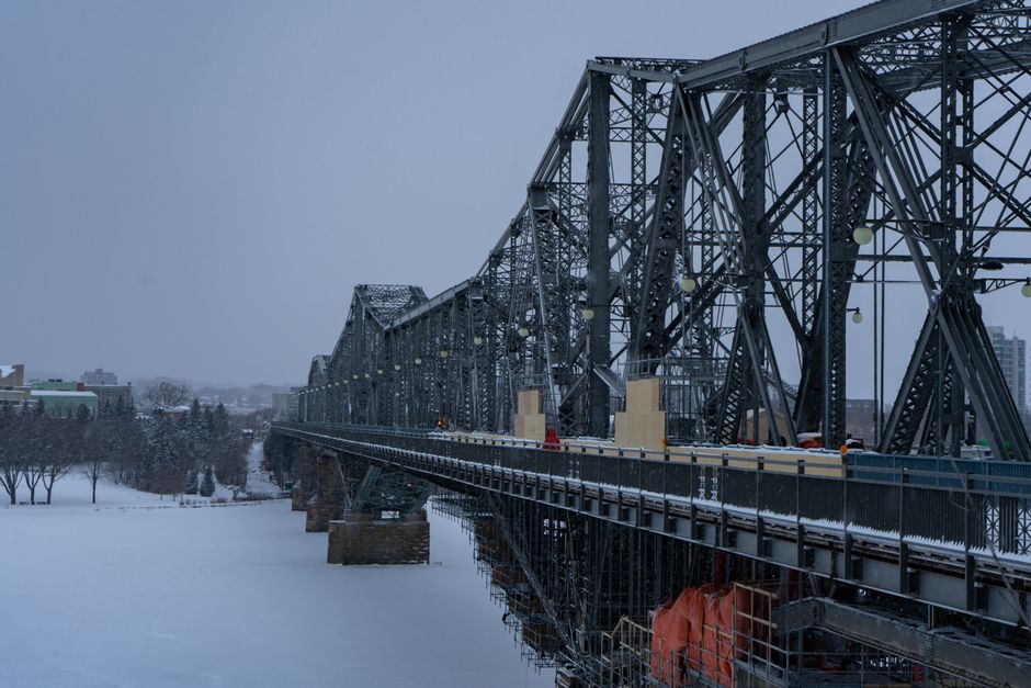 A black metal truss bridge with a lattice structure spans over a wide, frozen river during winter, with snow covering the ground and the bridge deck. The bridge features large support pillars beneath