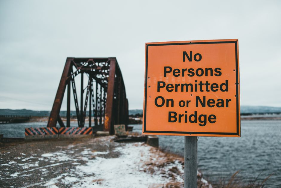 A black metal truss bridge with a lattice structure spans over a wide, frozen river during winter, with snow covering the ground and the bridge deck. The bridge features large support pillars beneath